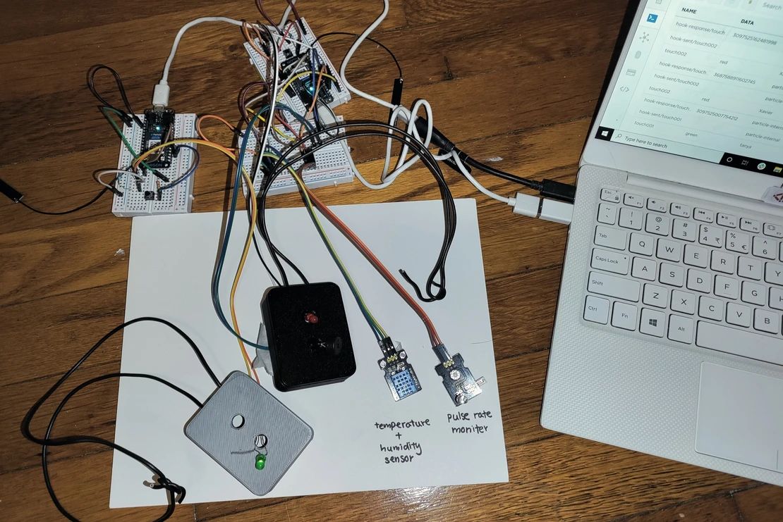 Hardware prototype showing Particle Argon microcontrollers on breadboards connected to temperature/humidity sensor and pulse rate monitor, with laptop showing Particle Cloud console